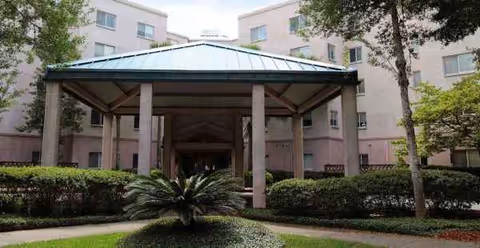Covered entrance pavilion with pillars sits before a landscaped courtyard and a beige multi-story building.
