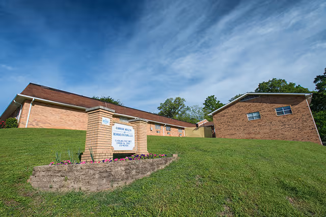 Exterior view of Cordova Health and Rehabilitation, LLC building on a grassy hill under a partly cloudy sky. The building is made of brick and has a sign in front surrounded by flowers.