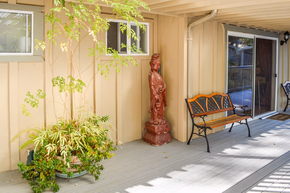 Outdoor covered patio area with a wooden bench with metal armrests, a large potted plant with green foliage, a red decorative statue, beige siding walls, and sliding glass doors leading inside.