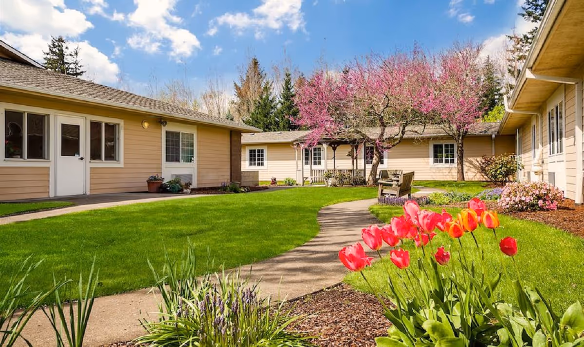 A bright outdoor courtyard area at The Cottages Senior Living Memory Care featuring a curved concrete pathway surrounded by green grass, blooming red and orange tulips, and pink flowering trees. Beige single-story buildings with white-trimmed windows and doors enclose the courtyard under a partly cloudy blue sky.