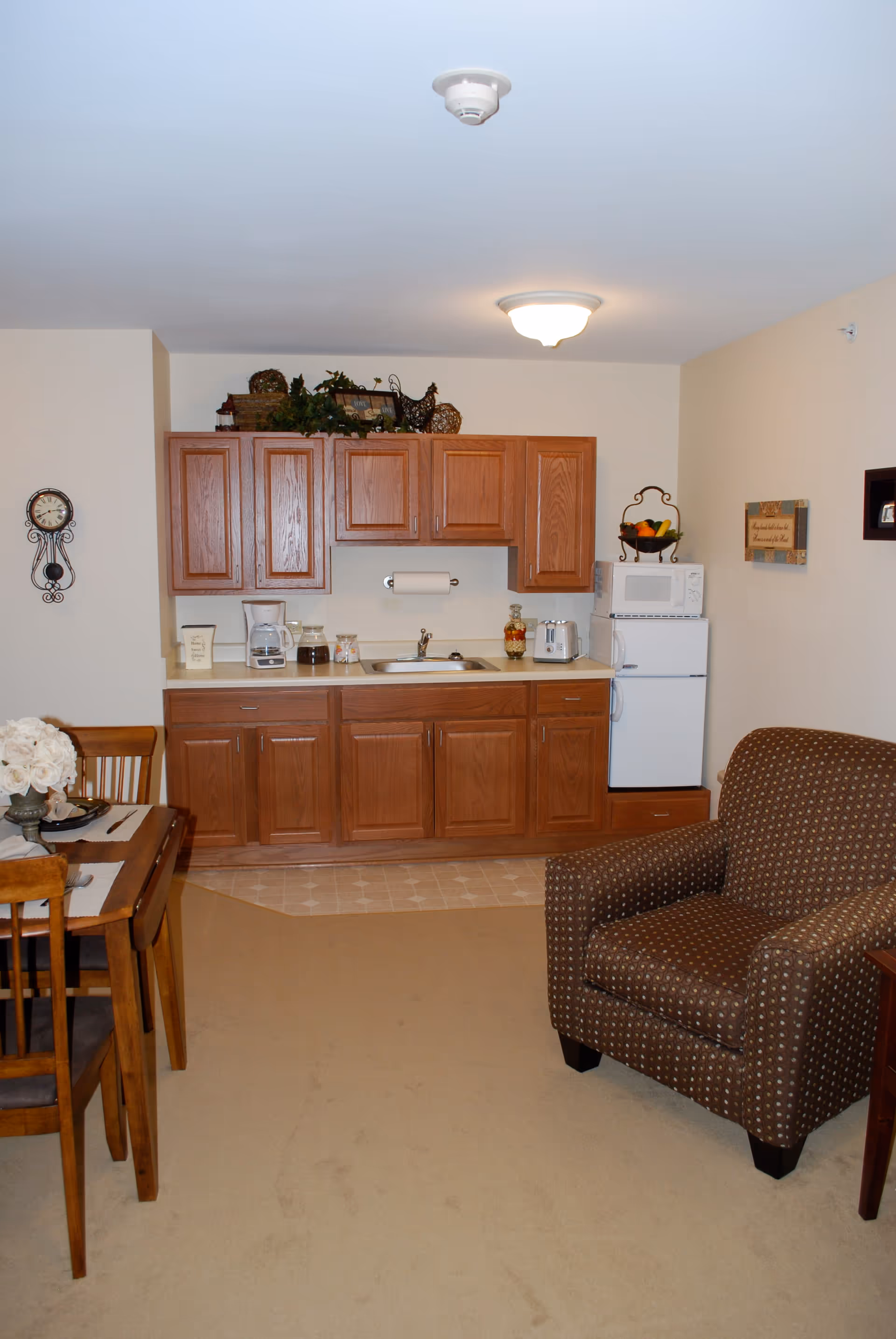 Small kitchenette in a senior living apartment with wooden cabinets, a sink, mini fridge and microwave, next to a dining table and upholstered chair.