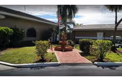 Front entrance courtyard of a mobile estates complex with a brick path leading to a central fountain and American flag, flanked by low buildings and palm trees.
