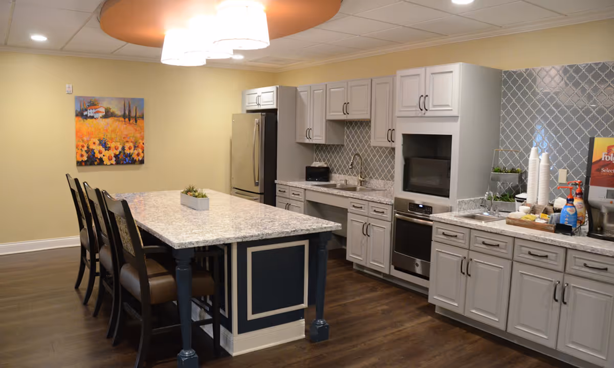 A modern kitchen area in a senior living facility featuring a large island with granite countertop and four high-back chairs. The kitchen has light gray cabinets, stainless steel appliances including a refrigerator and oven, a tiled backsplash, and a coffee station with cups and condiments. A colorful painting of a field of flowers hangs on the wall.