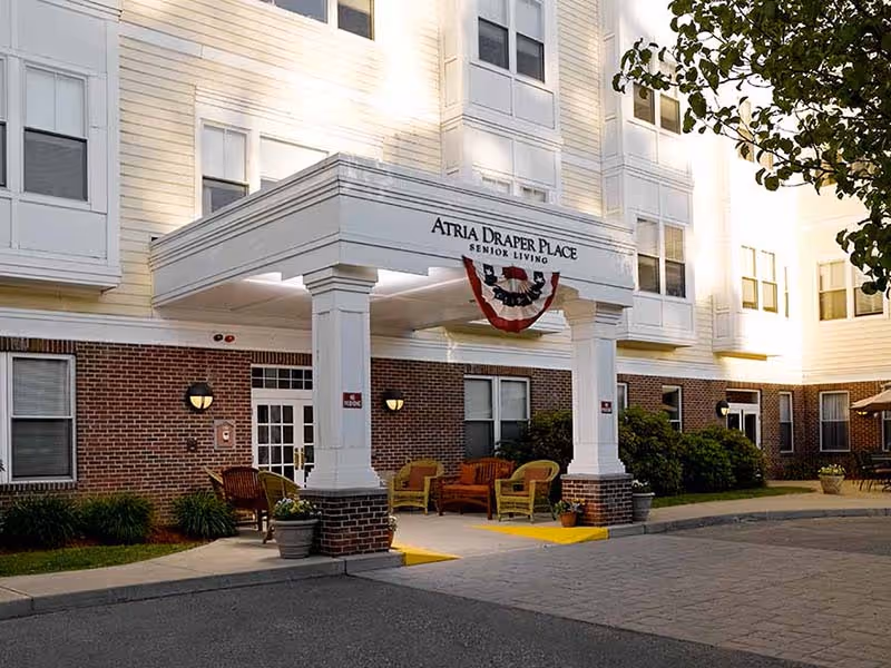 Entrance of Atria Draper Place senior living facility with a covered drop-off area supported by white columns. There are outdoor chairs and tables arranged near the entrance, with brick and white siding on the building exterior. A patriotic bunting decoration hangs above the entrance.