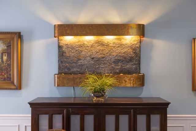 A decorative indoor water feature mounted on a light blue wall above a dark wooden cabinet. The water feature has a textured stone surface with a copper frame and is illuminated by three small lights at the top. A small green plant in a glass pot is placed on the cabinet below the water feature. Two framed paintings are partially visible on either side of the water feature.