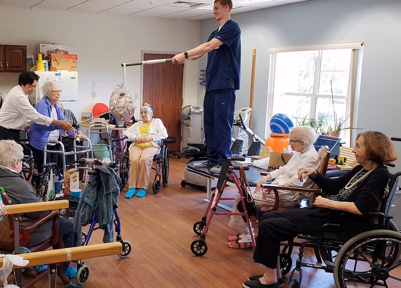 A group of elderly individuals, some in wheelchairs and others using walkers, participate in a recreational activity in a room with wooden flooring and exercise equipment. A young staff member in blue scrubs stands on a platform holding a stick to hit a hanging piñata, while another staff member assists an elderly woman with a stick. The room has a window letting in natural light and various exercise balls and equipment in the background.