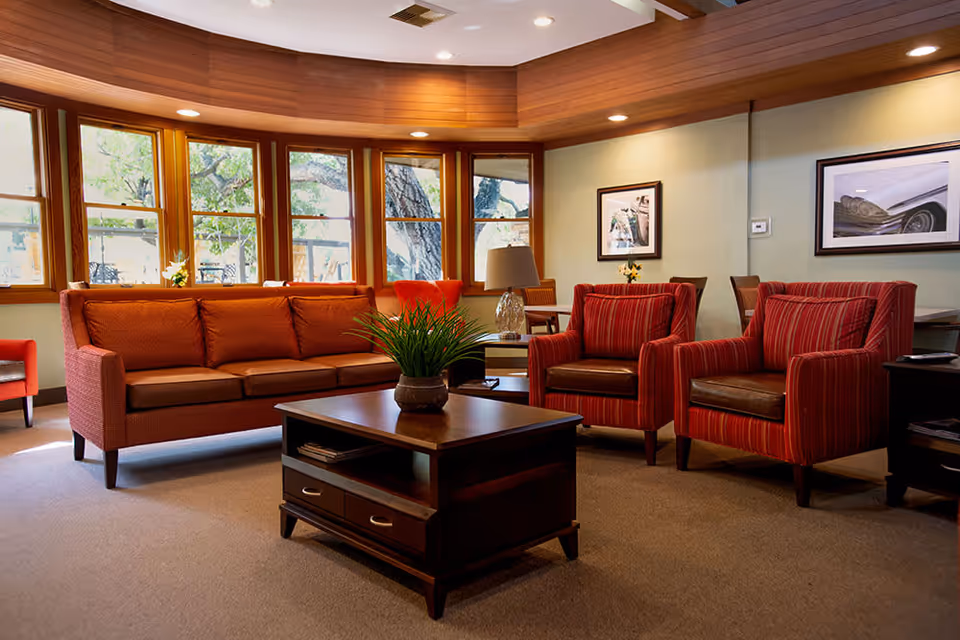 A cozy living room area with a brown sofa, two red striped armchairs, a wooden coffee table with a potted plant, and large windows letting in natural light. The walls have framed pictures and there is a table lamp on a side table.