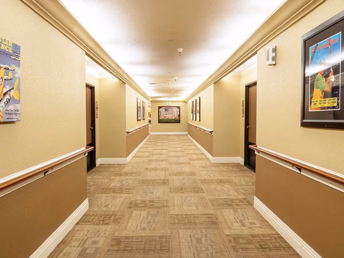 A well-lit hallway in an assisted living facility with beige walls and carpeted floor. The hallway has handrails on both sides and several closed doors along the walls. Framed vintage-style posters are hung on the walls, and an exit sign is visible at the far end above a door.