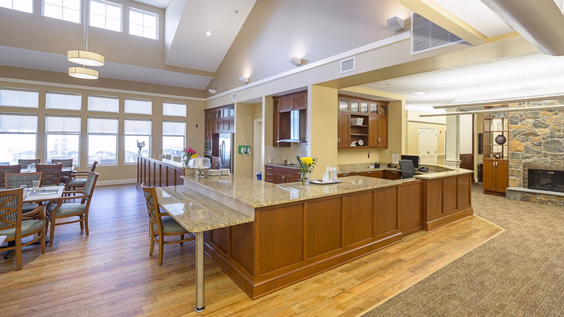 A spacious and well-lit dining area and kitchen in a senior living facility. The kitchen features wooden cabinets, granite countertops, and modern appliances. Adjacent to the kitchen is a dining area with wooden tables and chairs, large windows letting in natural light, and a high ceiling with hanging light fixtures. There is also a stone fireplace and a carpeted area visible in the background.