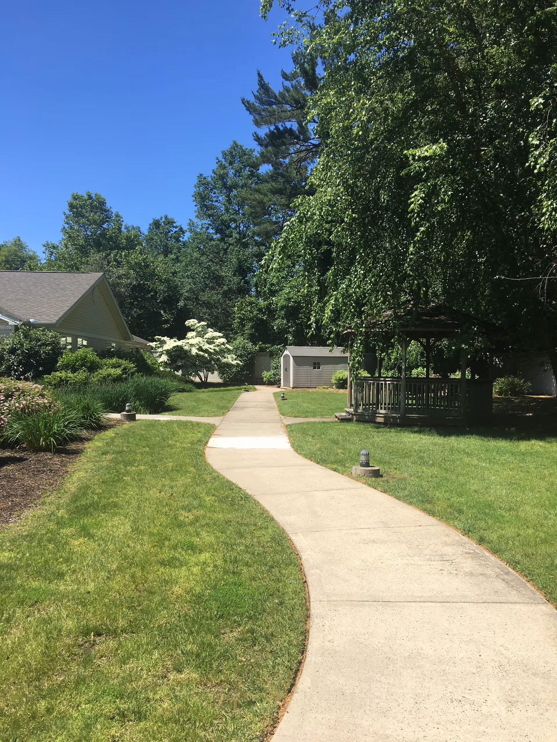 A winding concrete pathway through a well-maintained green lawn with trees and bushes on either side. There is a small gazebo on the right side and a shed in the background. The sky is clear and blue.