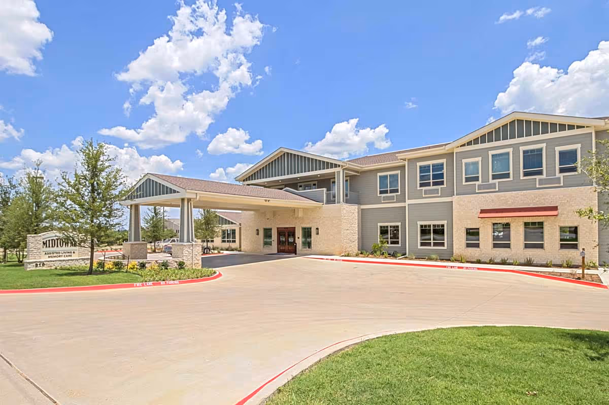 Exterior view of Midtowne Assisted Living and Memory Care building under a partly cloudy blue sky, showing a two-story structure with a covered entrance driveway, landscaped greenery, and a sign with the facility name.