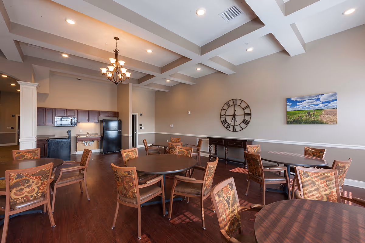 A spacious dining room with several round tables and patterned cushioned chairs. The room features a coffered ceiling with recessed lighting and a chandelier. There is a kitchenette area with dark wood cabinets, a black refrigerator, and a microwave. On the wall, there is a large decorative clock and a landscape painting. The floor is dark wood, and the walls are painted in neutral tones.