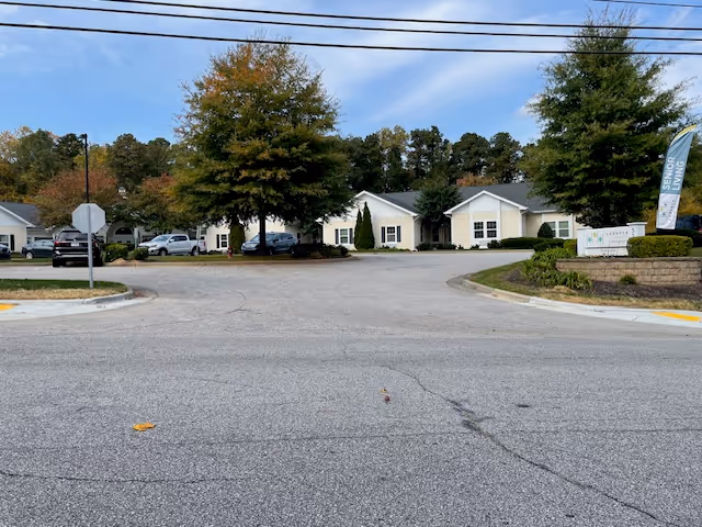 View of the entrance to Cadence North Raleigh senior living facility showing a paved driveway, several parked cars, trees with autumn foliage, and a sign with the facility's name and a flag that reads 'Senior Living'.