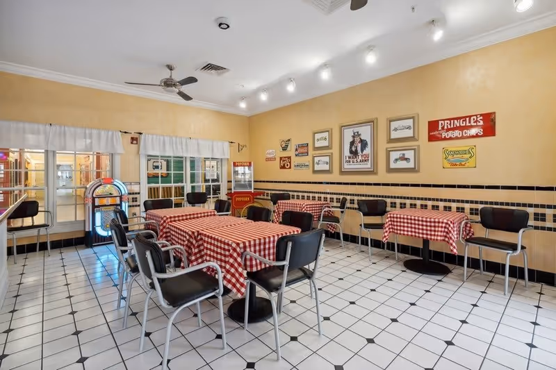 A dining area with several tables covered in red and white checkered tablecloths surrounded by black chairs. The walls are decorated with vintage-style signs and framed pictures. There is a popcorn machine and a jukebox near the windows, and the floor is tiled with a white and black pattern.
