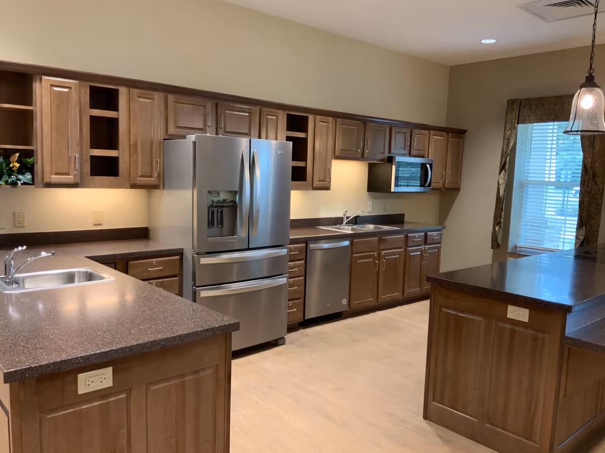 A spacious kitchen with wooden cabinets, a stainless steel refrigerator, dishwasher, microwave, and two sinks. The countertops are dark brown, and there is a window with curtains letting in natural light. The floor is light-colored wood, and there are pendant lights hanging from the ceiling.