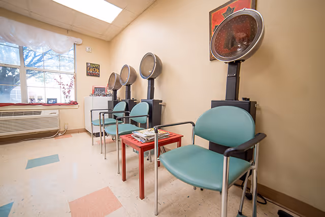 Interior view of a hair salon area in a senior living facility with four green salon chairs each positioned in front of hair drying machines. A small red table with magazines is placed between the chairs. A large window with white curtains and an air conditioning unit is visible on the left side of the room.