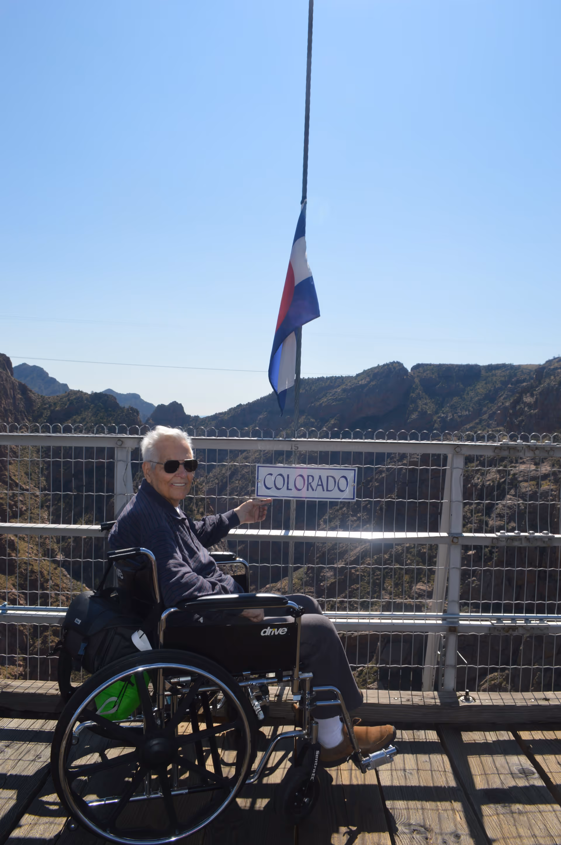 An elderly man in a wheelchair wearing sunglasses and a dark jacket is sitting on a wooden platform with a metal railing. He is smiling and pointing at a sign that says 'COLORADO'. Behind him, there is a mountainous landscape under a clear blue sky, and a flag is hanging on a pole above the sign.