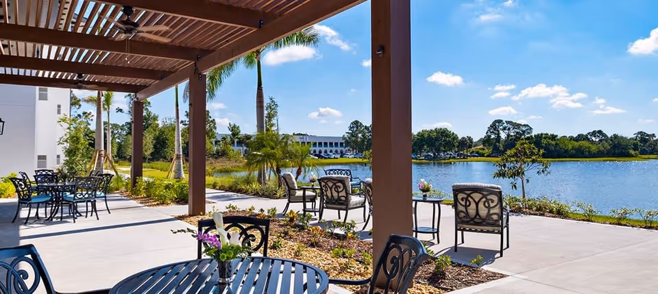 Outdoor patio area with metal tables and chairs under a wooden pergola, overlooking a lake with trees and a building in the background under a blue sky with scattered clouds.