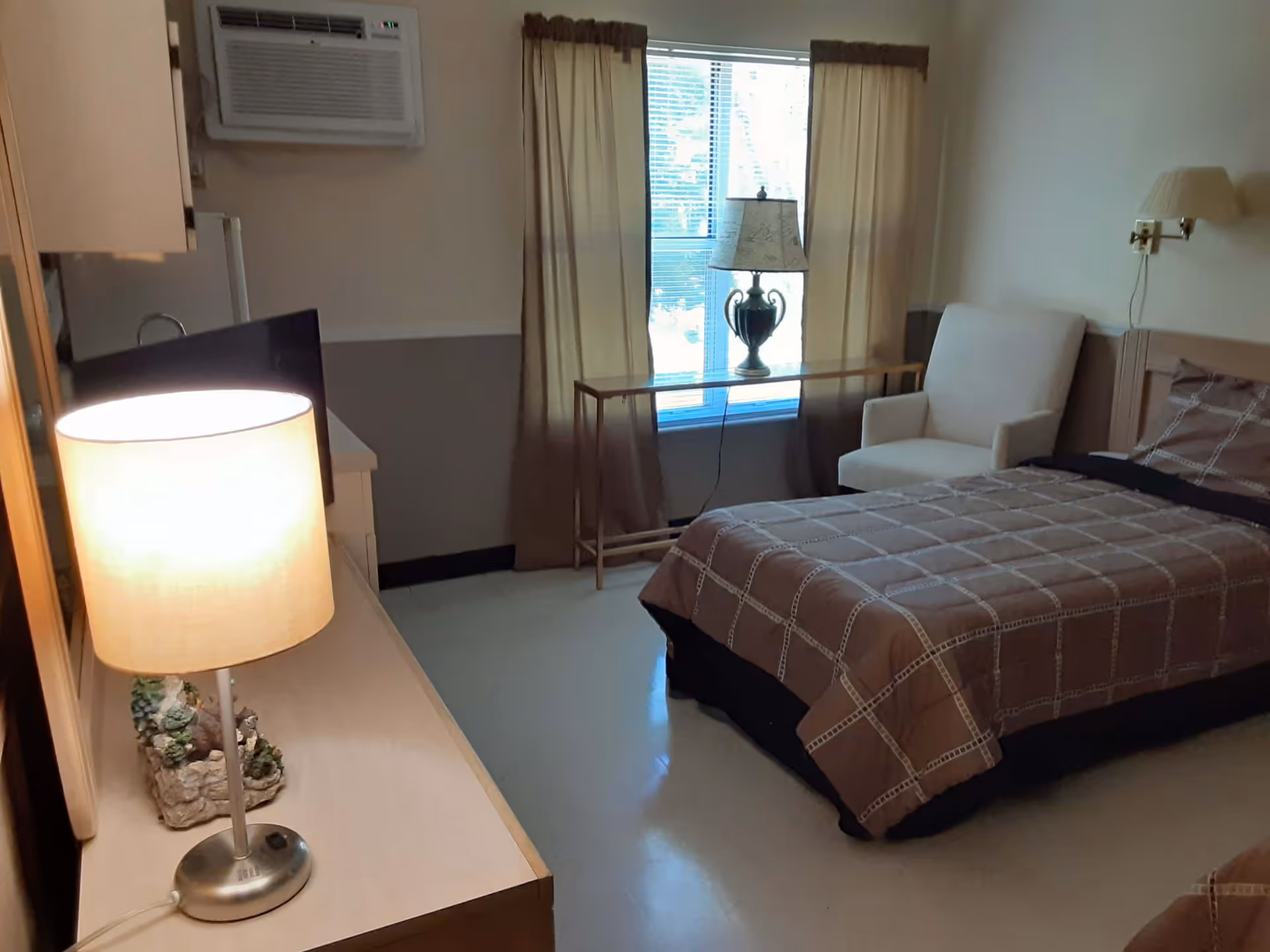 A small bedroom with a single bed covered in a brown checkered bedspread, a beige armchair, a window with beige curtains, a table with a decorative lamp, a wall-mounted air conditioner, and a dresser with a lit table lamp and a small decorative item.
