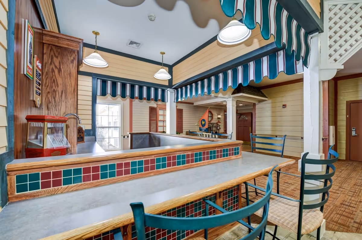 Interior view of a communal dining or snack area with a counter featuring red and green tiles, high chairs with metal frames and woven seats, striped fabric awnings overhead, pendant lights, and a popcorn machine on the left side. The walls are paneled with light-colored wood, and there are windows and doors in the background.