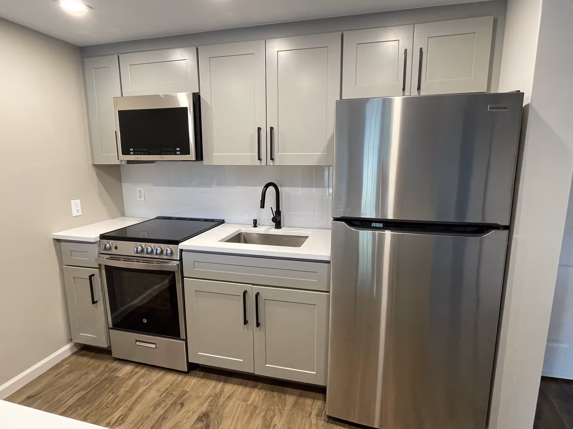 Modern kitchen area with white cabinets, a stainless steel refrigerator, a stove with an oven, a microwave above the stove, a sink with a black faucet, and wood-style flooring.