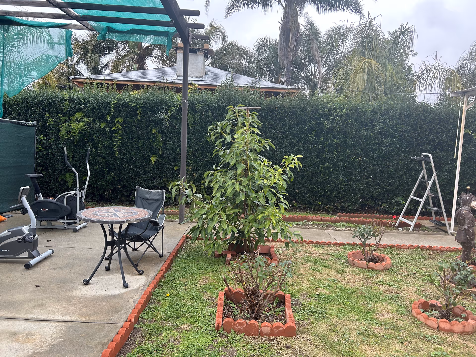 Outdoor garden area with a small patio featuring a round mosaic table and a folding chair. There are exercise machines on the concrete patio, a green hedge fence in the background, a small tree and several flower beds bordered with red bricks. A metal ladder and a statue are also visible in the garden.