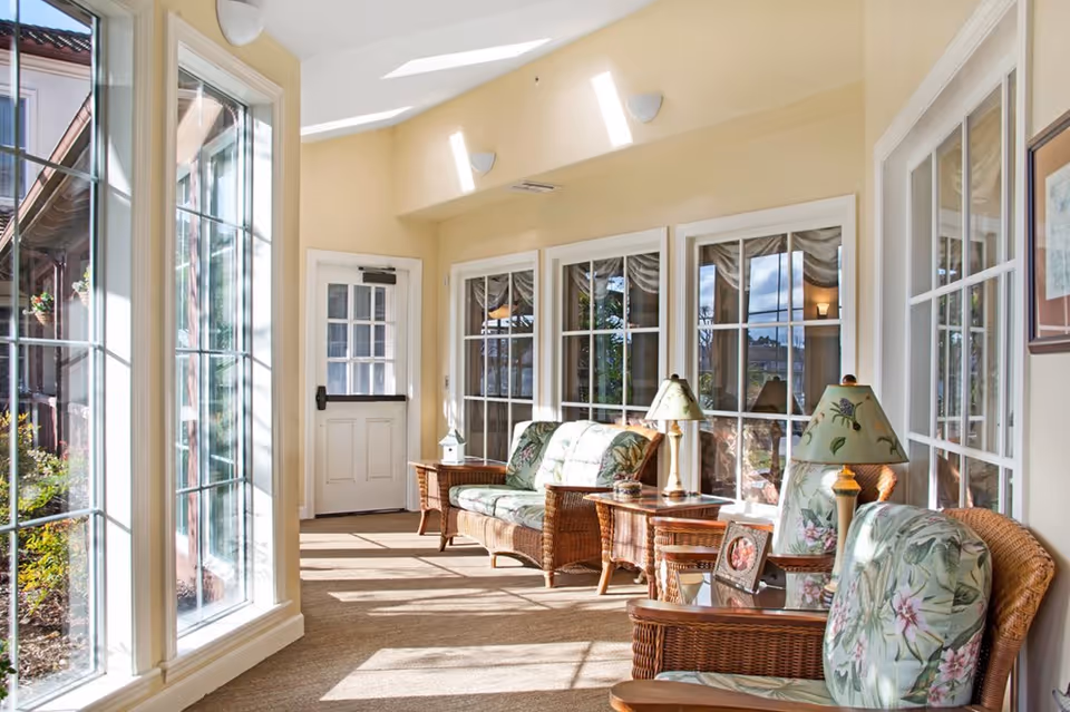 Sunlit sunroom with wicker seating, floral cushions, side tables and lamps along large windows and a glass door.