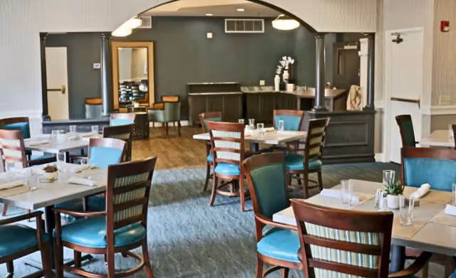 A dining room in a senior living facility with multiple tables and chairs arranged neatly. The chairs have wooden frames with teal cushions. Tables are set with glasses, napkins, and small decorative plants. The room features a carpeted floor, a large mirror on the back wall, and soft lighting from ceiling fixtures.