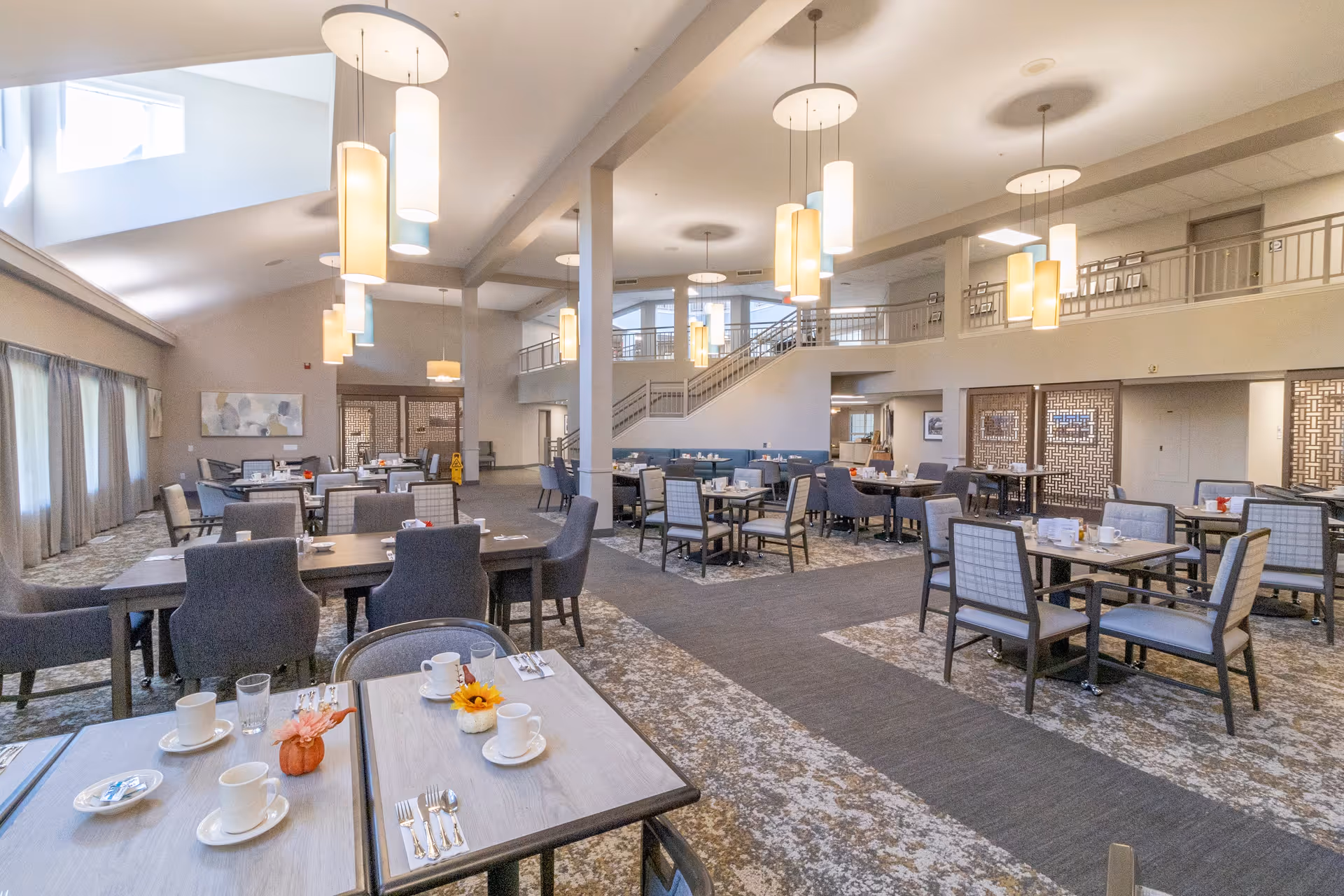 Spacious dining room in a senior living facility with multiple tables and chairs arranged neatly. The room features modern pendant lighting, large windows with curtains, and a high ceiling with a mezzanine level. Tables are set with cups, plates, and small decorative items.
