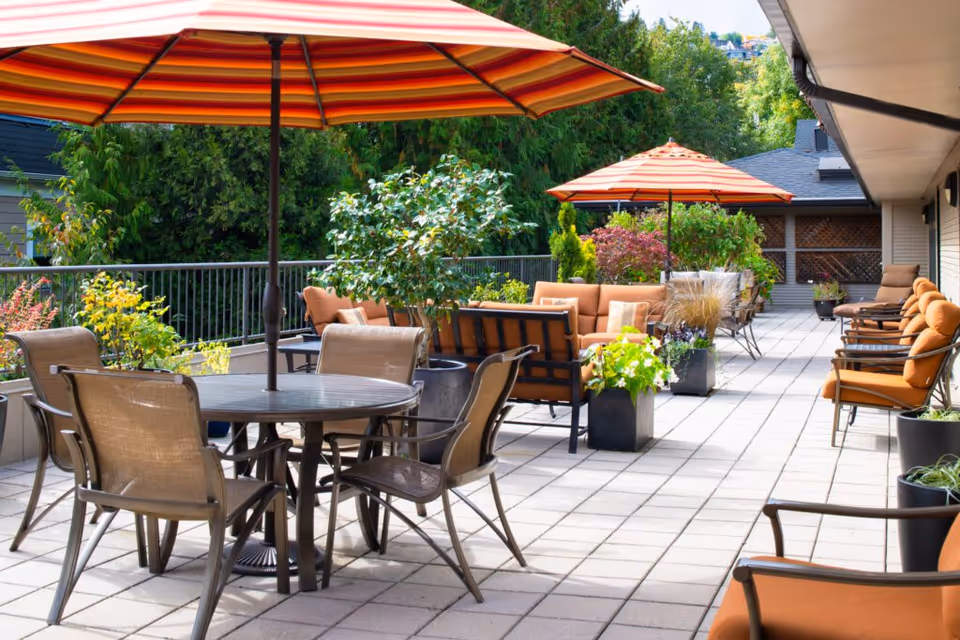 Outdoor patio area with multiple seating arrangements including round tables with chairs and orange cushioned sofas under large orange striped umbrellas, surrounded by potted plants and greenery.