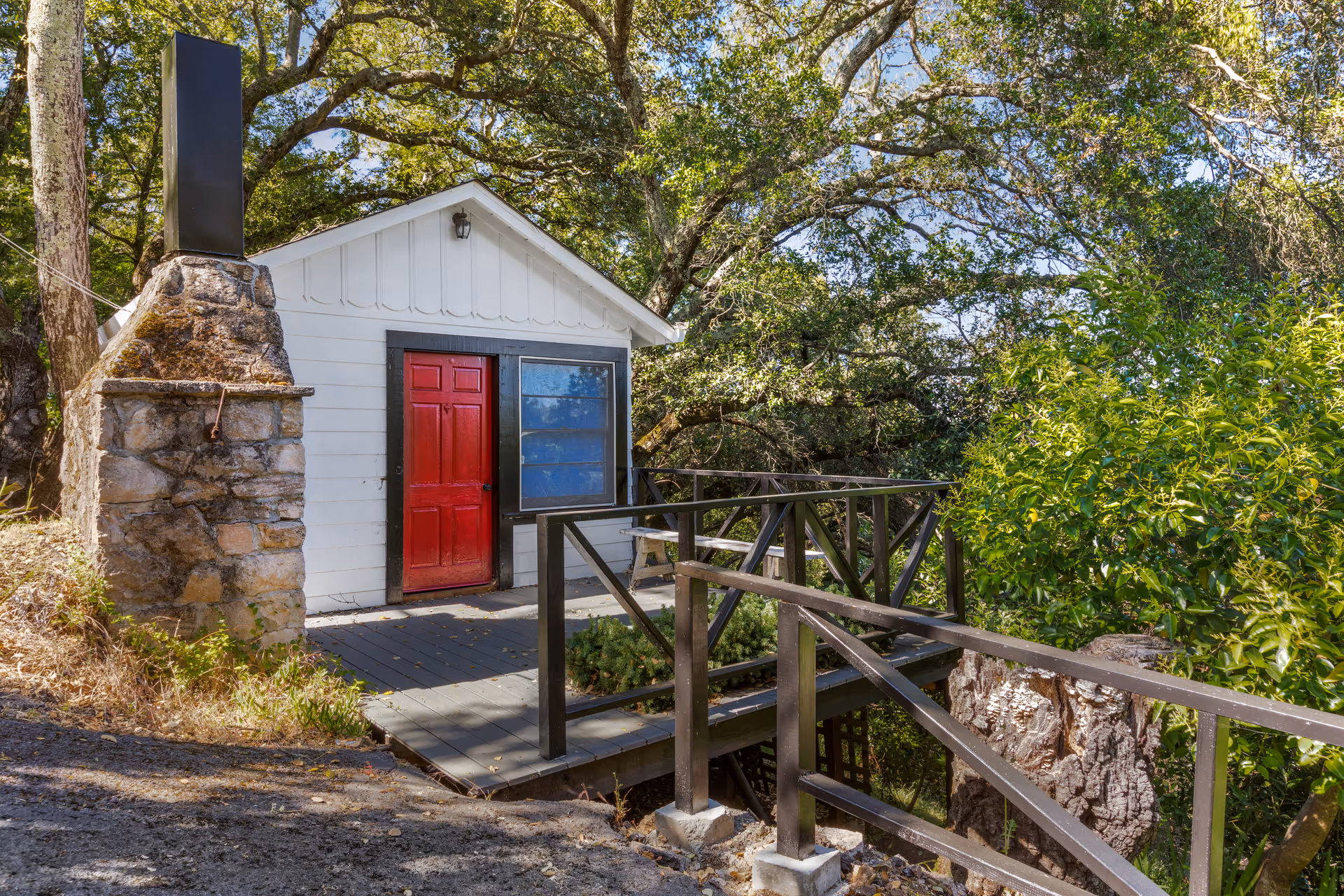 Small white building with a bright red door and a large window, surrounded by trees and greenery. There is a wooden ramp with railings leading up to the entrance, and a stone chimney structure on the left side. The scene is outdoors with natural sunlight filtering through the trees.