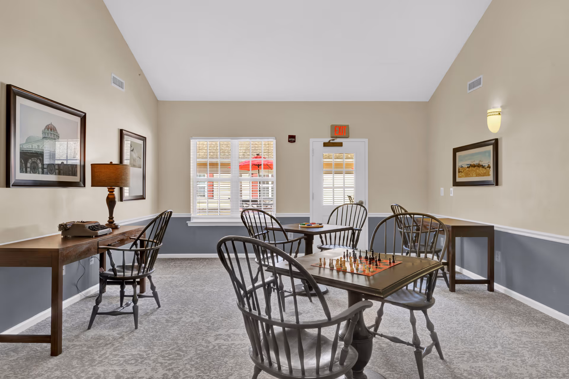 A well-lit activity room with beige and gray walls, featuring several wooden tables and chairs. One table has a chessboard set up, and another table has a board game. There are framed pictures on the walls, a lamp on a side table with a vintage typewriter, and a window and door with glass panes letting in natural light.