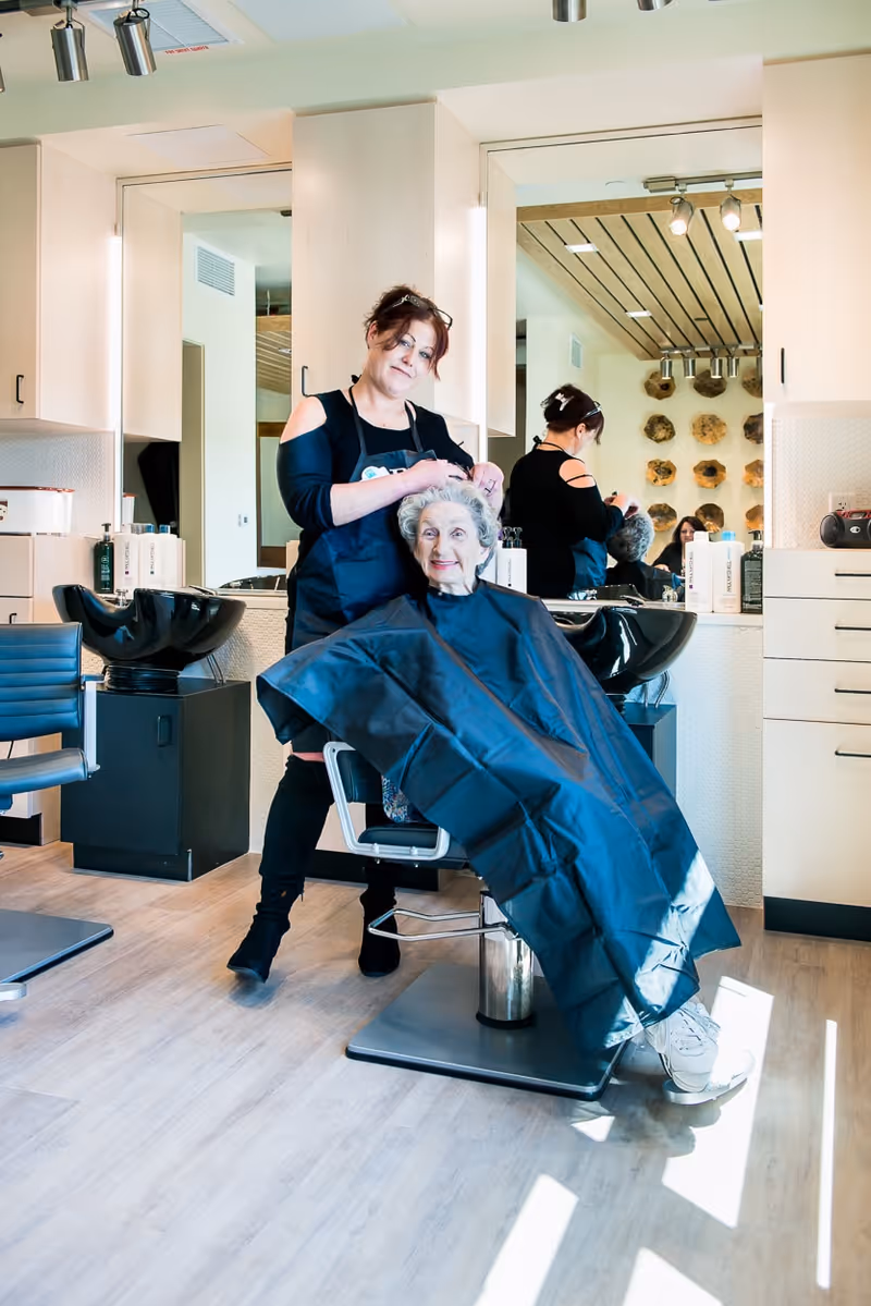A hairdresser styling the hair of an elderly woman seated in a salon chair, both smiling. The salon has light-colored walls, large mirrors, and modern hair washing stations in the background.