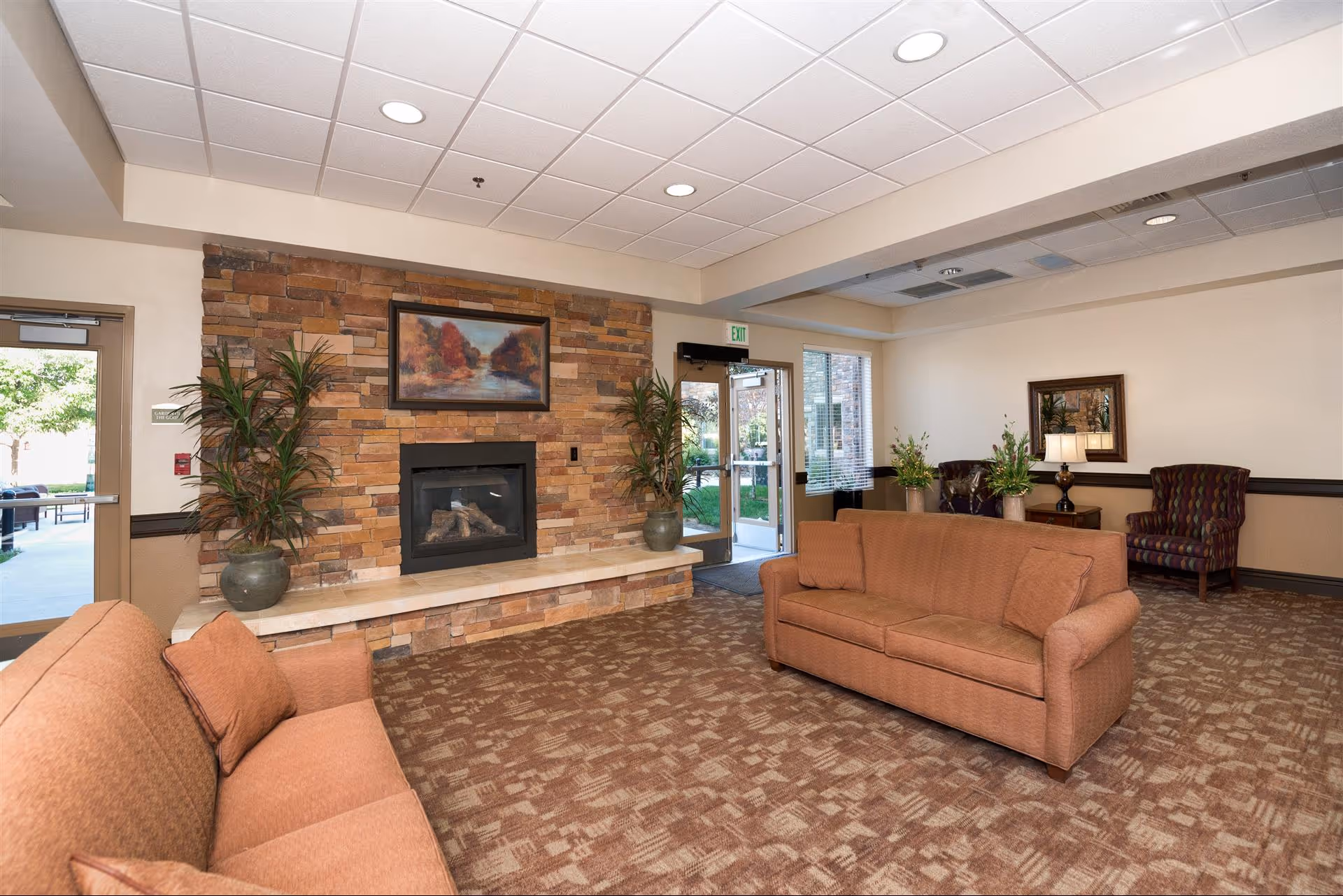 A cozy living room area in Bethesda Gardens featuring two orange sofas, a stone fireplace with a landscape painting above it, two large potted plants on either side of the fireplace, patterned carpet, and a side table with a lamp and floral arrangements. There are glass doors leading outside and a window letting in natural light.