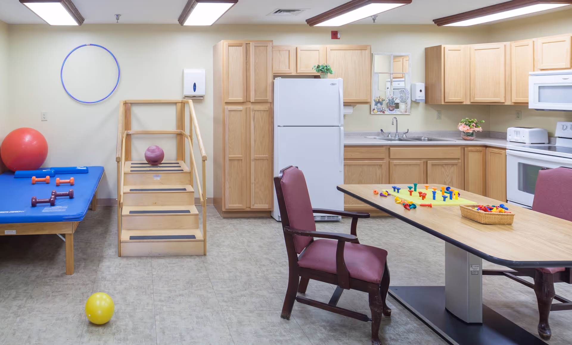 A room with a kitchen area featuring wooden cabinets, a white refrigerator, microwave, stove, and sink. In the foreground, there is a table with two chairs and colorful pegboard games on the table. To the left, there is a blue therapy table with small dumbbells, a set of wooden stairs with handrails, a red exercise ball, a yellow ball on the floor, and a blue hula hoop hanging on the wall.