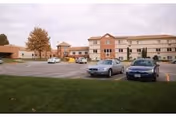 Front view of a multi-story senior living building with a parking lot and several parked cars under an overcast sky.