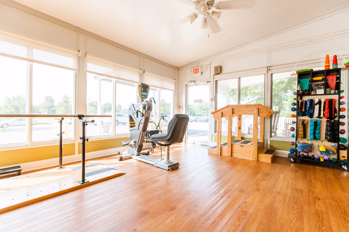 Bright rehabilitation exercise room with parallel bars, a recumbent exercise bike, a wooden therapy platform and a rack of therapy equipment by large windows.