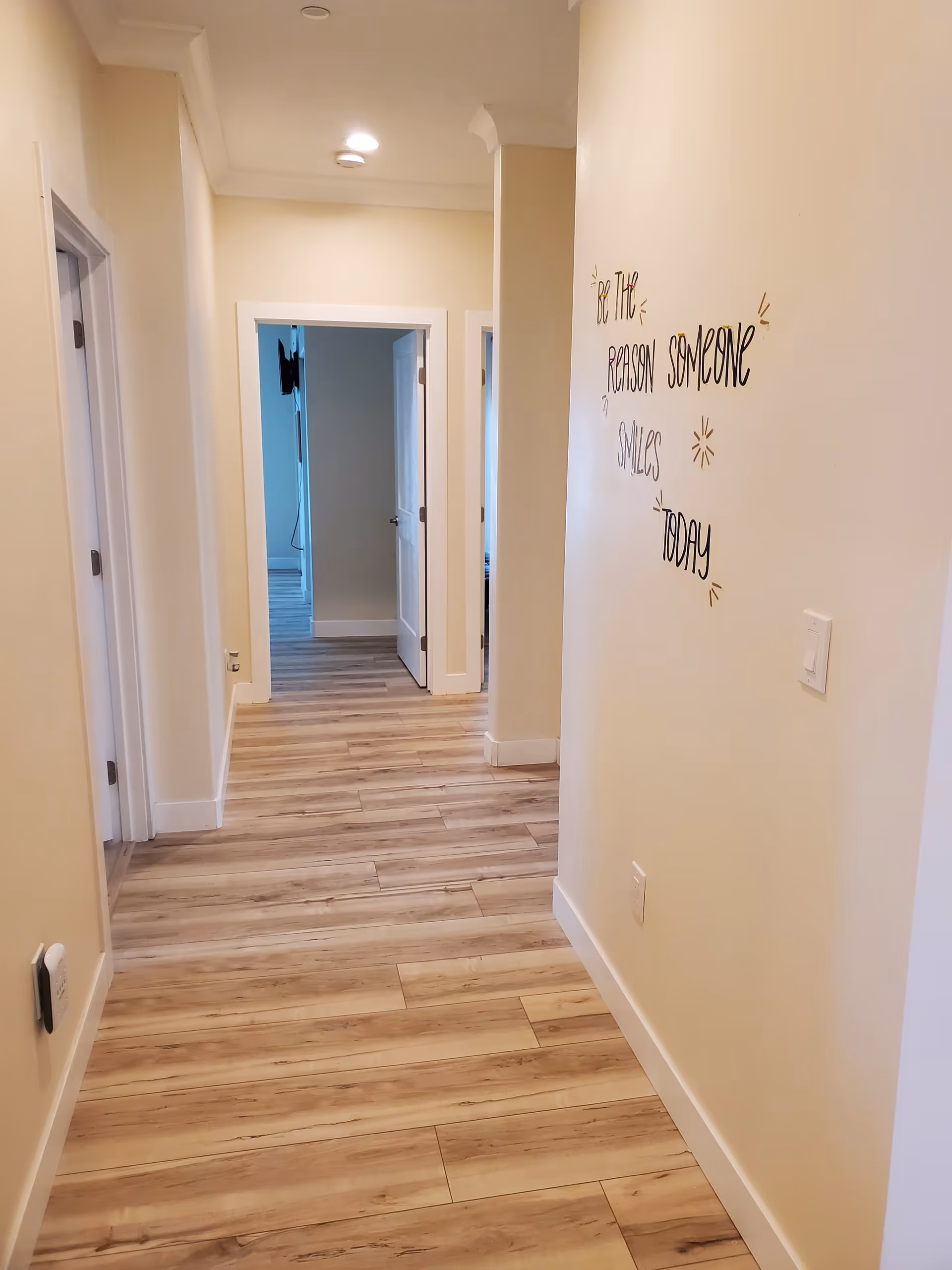 Hallway with wood-look floors, cream walls, recessed ceiling lights, several open doorways, and a motivational wall decal.