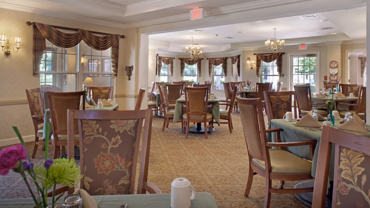 A spacious dining room with multiple tables covered in green tablecloths, each set with napkins, cups, and small flower arrangements. The room features large windows with decorative curtains, beige walls, carpeted floors, and elegant chandeliers hanging from the ceiling.