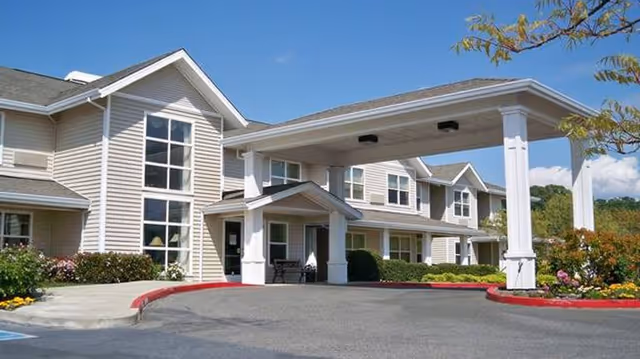Exterior view of a senior living facility building with beige siding, large windows, and a covered entrance driveway. The building is surrounded by landscaped bushes and flowers under a clear blue sky.