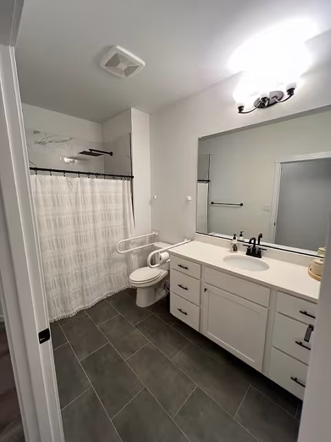 A clean bathroom with a white vanity and sink, a large mirror above the sink, a toilet with a safety grab bar, and a shower with a white curtain and dark shower rod. The floor is covered with dark gray tiles and the walls are painted white. There is a ceiling vent and a light fixture above the mirror.