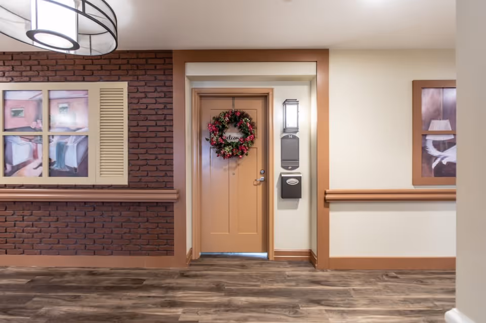 Interior hallway of a senior living facility with a closed door decorated with a floral wreath and a welcome sign. The door is framed by a light fixture, a mailbox, and a room number plaque. The hallway features wood flooring, brick and beige walls, and framed artwork on either side of the door.
