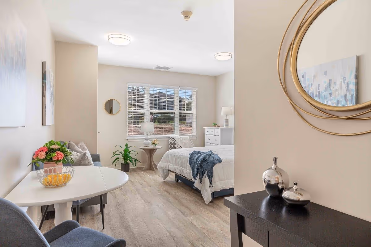 A bright and airy bedroom in a senior living facility featuring a bed with white bedding and a blue throw, a white dresser, a small round side table with a lamp and flowers, a window with white blinds, a small dining table with chairs, a plant, and decorative wall art including a round mirror and a painting.