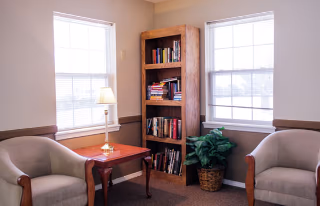 A cozy corner of a living room or common area with two beige armchairs facing each other, a wooden side table with a lamp, a tall wooden bookshelf filled with books, and a potted plant next to the bookshelf. Two large windows with blinds allow natural light into the room.
