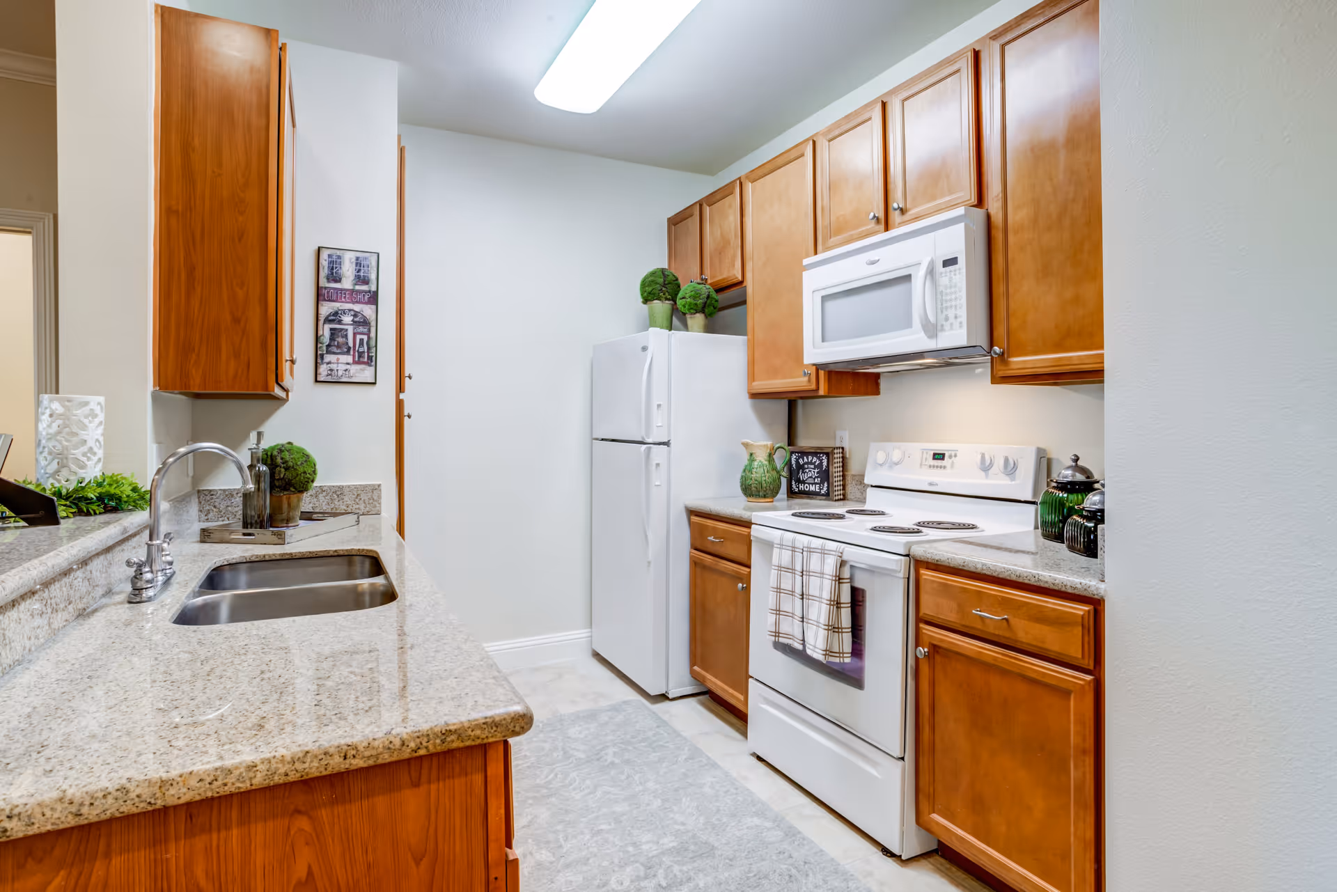 A clean and modern kitchen with wooden cabinets, a white refrigerator, white microwave, and white electric stove. The countertop is granite with a double sink and a silver faucet. There are decorative plants and kitchen items on the counters, and a framed picture on the wall.
