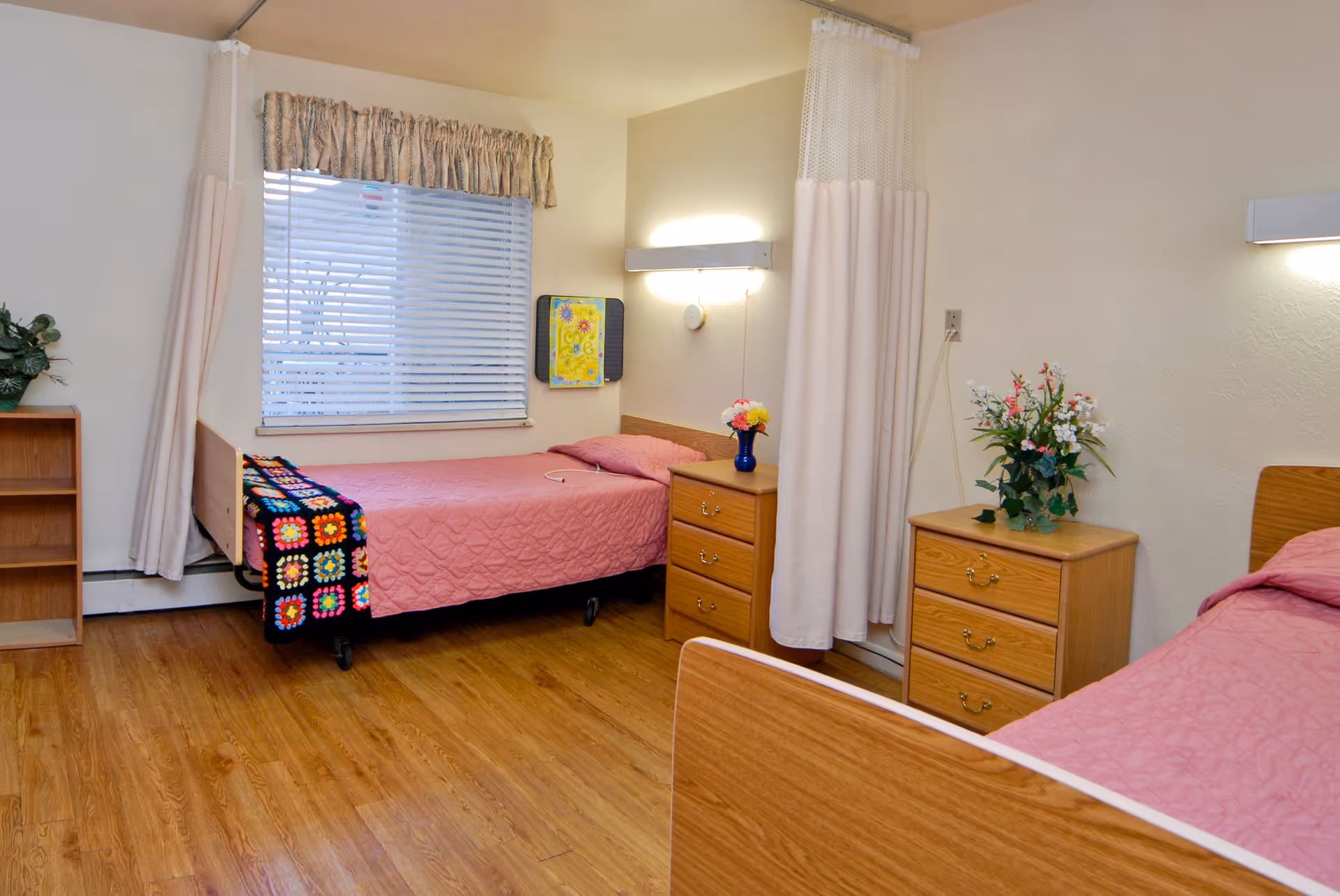 A clean and simple senior care facility bedroom with two single beds, each covered with a pink quilt. There are wooden nightstands with drawers next to each bed, one with a vase of flowers. A window with blinds and a floral valance is on the wall, and a privacy curtain is partially drawn between the beds. The floor is wood, and the walls are light-colored.