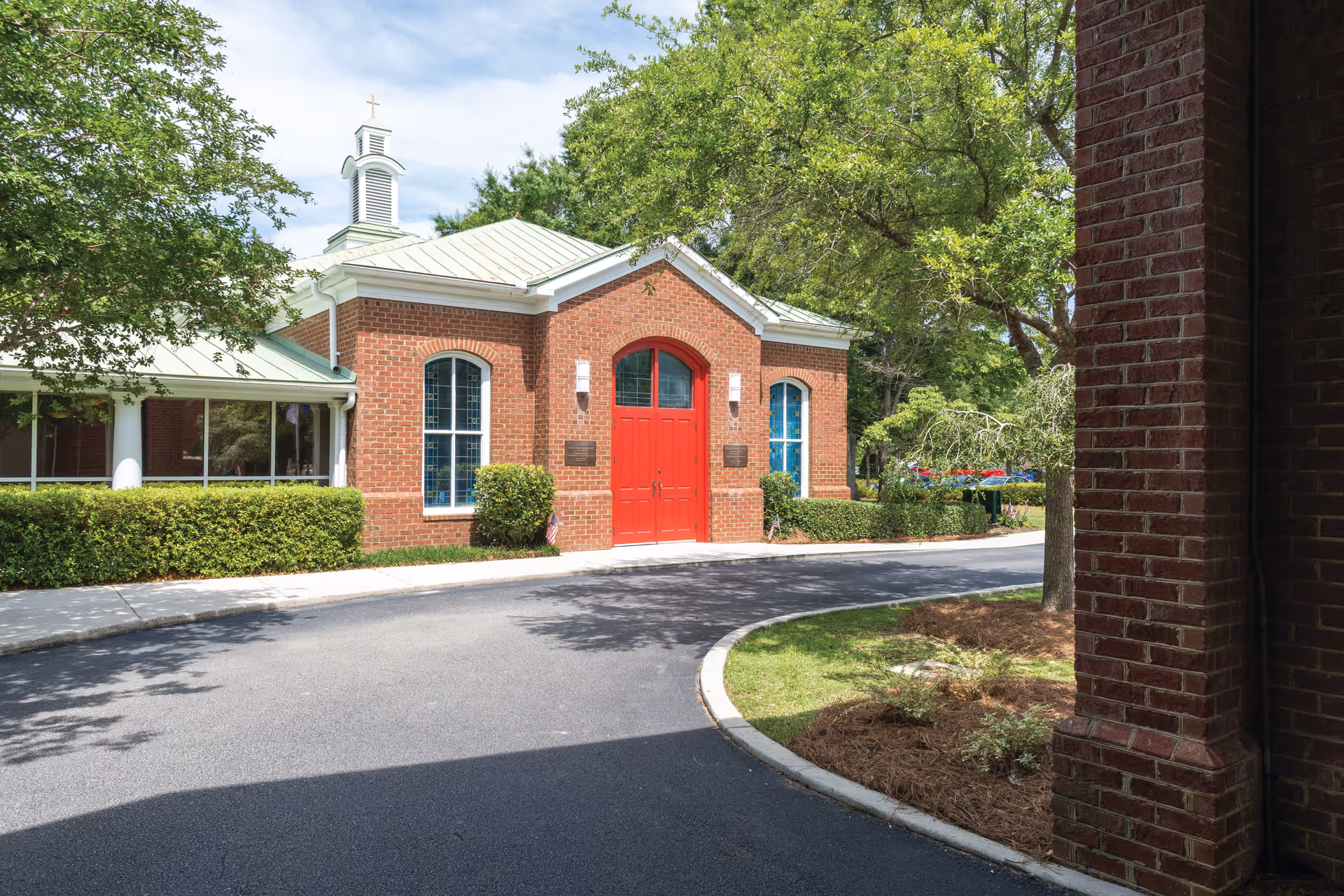Exterior view of a brick building with a green metal roof and a small cupola topped with a cross. The building has a prominent red double door entrance and arched windows. It is surrounded by trees, bushes, and a curved driveway.