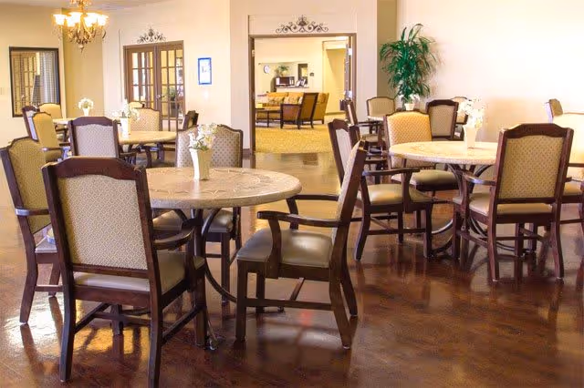 A well-lit dining area with multiple round tables and cushioned wooden chairs arranged neatly. Each table has a small vase with white flowers. The room features polished wooden floors, a chandelier, and a large potted plant in the corner. In the background, there is an open doorway leading to another room with additional seating.