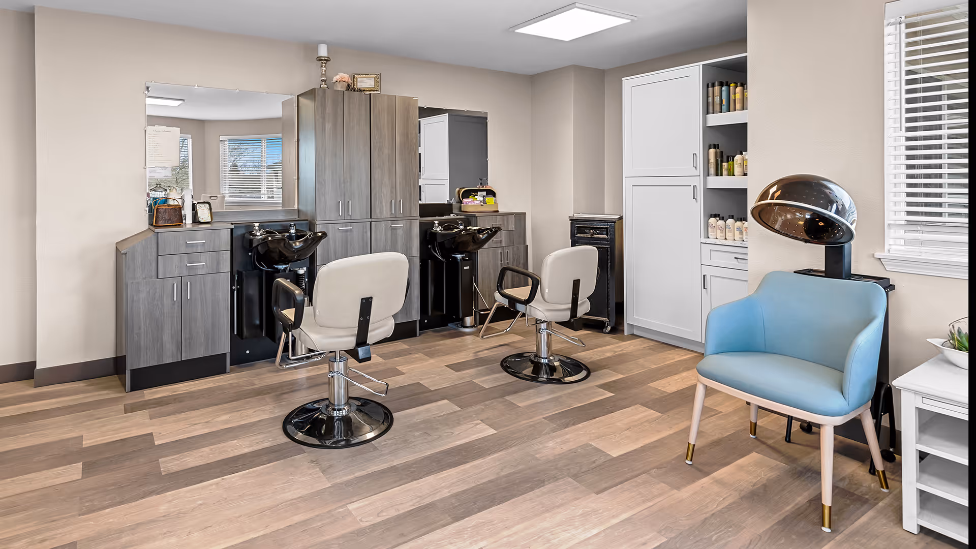 Interior view of a hair salon area in a senior living facility with two salon chairs facing mirrors and wash basins, wooden cabinets for storage, a blue waiting chair, and a hair dryer near a window with blinds.