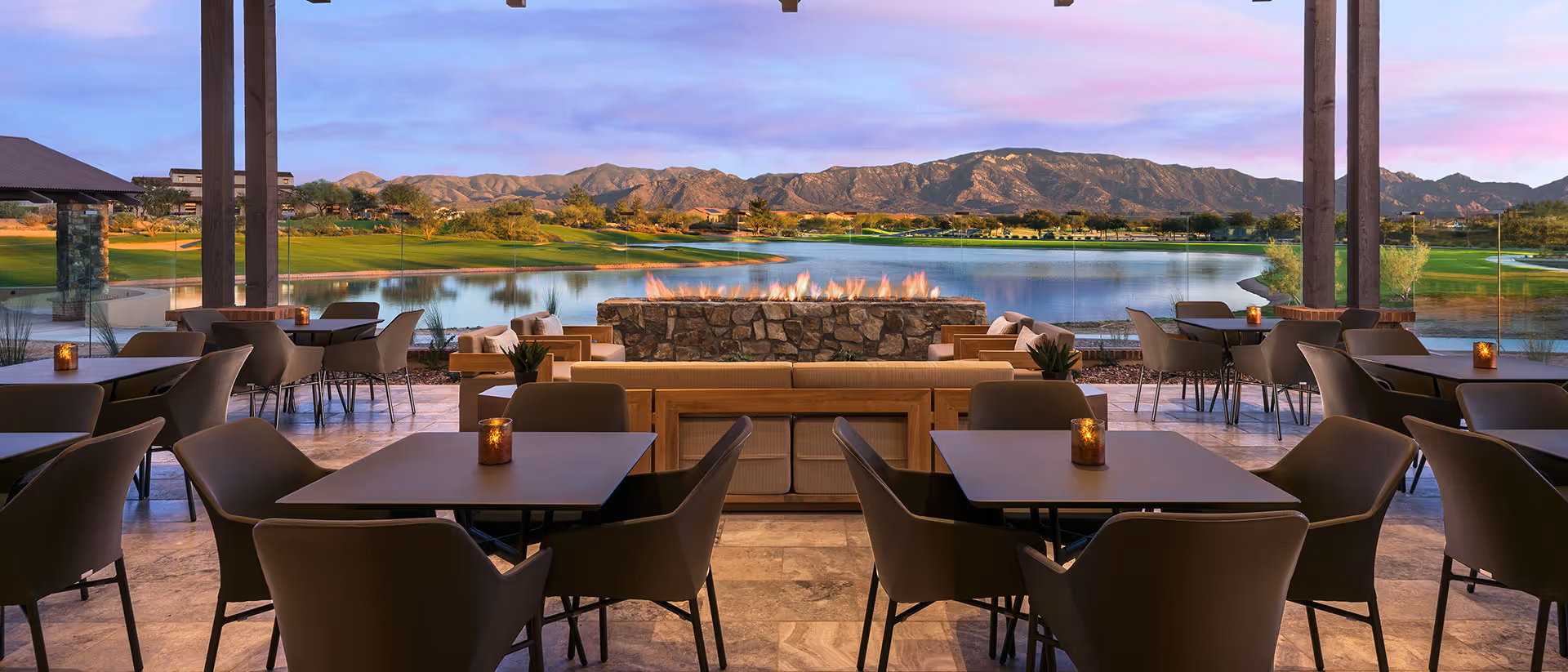 Covered outdoor dining terrace with tables and chairs facing a stone fire pit, lake, and mountain view at sunset.